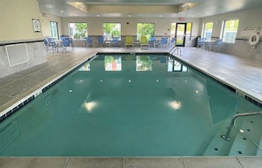 Una piscina interior con agua cristalina, rodeada de sillas y mesas azules, y grandes ventanales que dejan entrar luz natural.