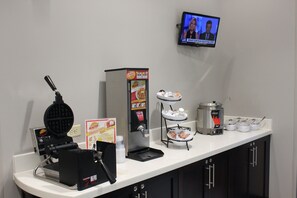 A self-service breakfast station with a waffle maker, coffee machine, and a TV displaying news.