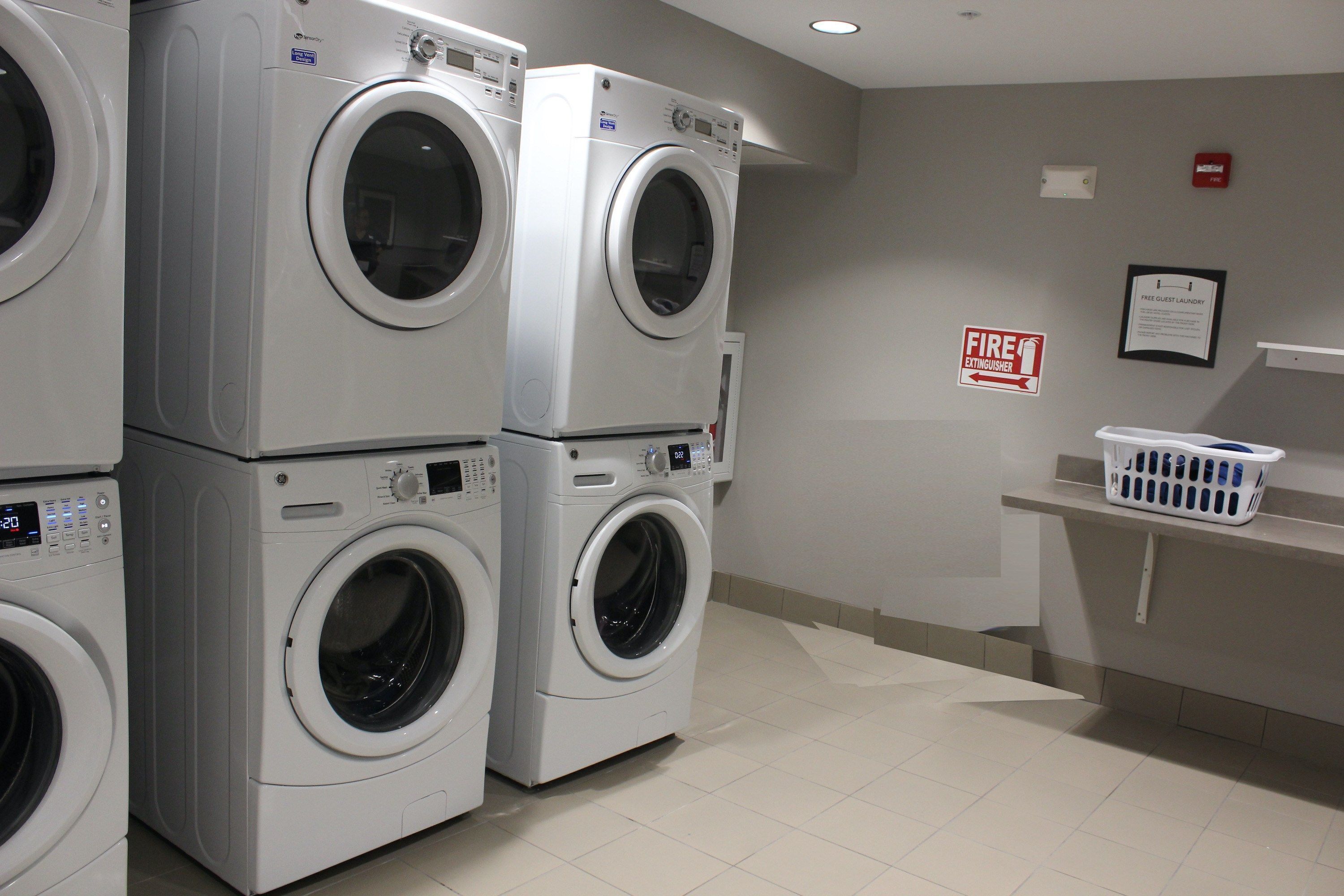 A laundry room with stacked front-loading washers and dryers, a fire extinguisher sign, and a notice about free guest laundry.