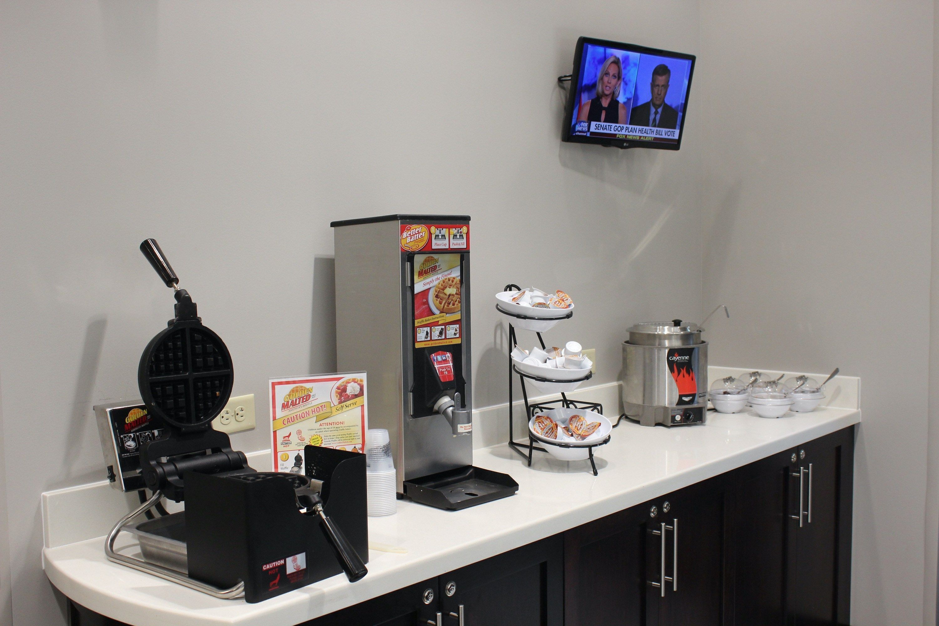 A self-service breakfast station with a waffle maker, coffee machine, and a TV displaying news.