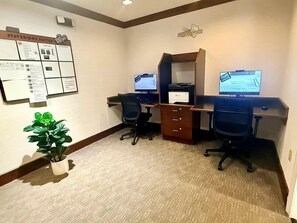 A hotel room with two desks, a potted plant, and a bulletin board with information.