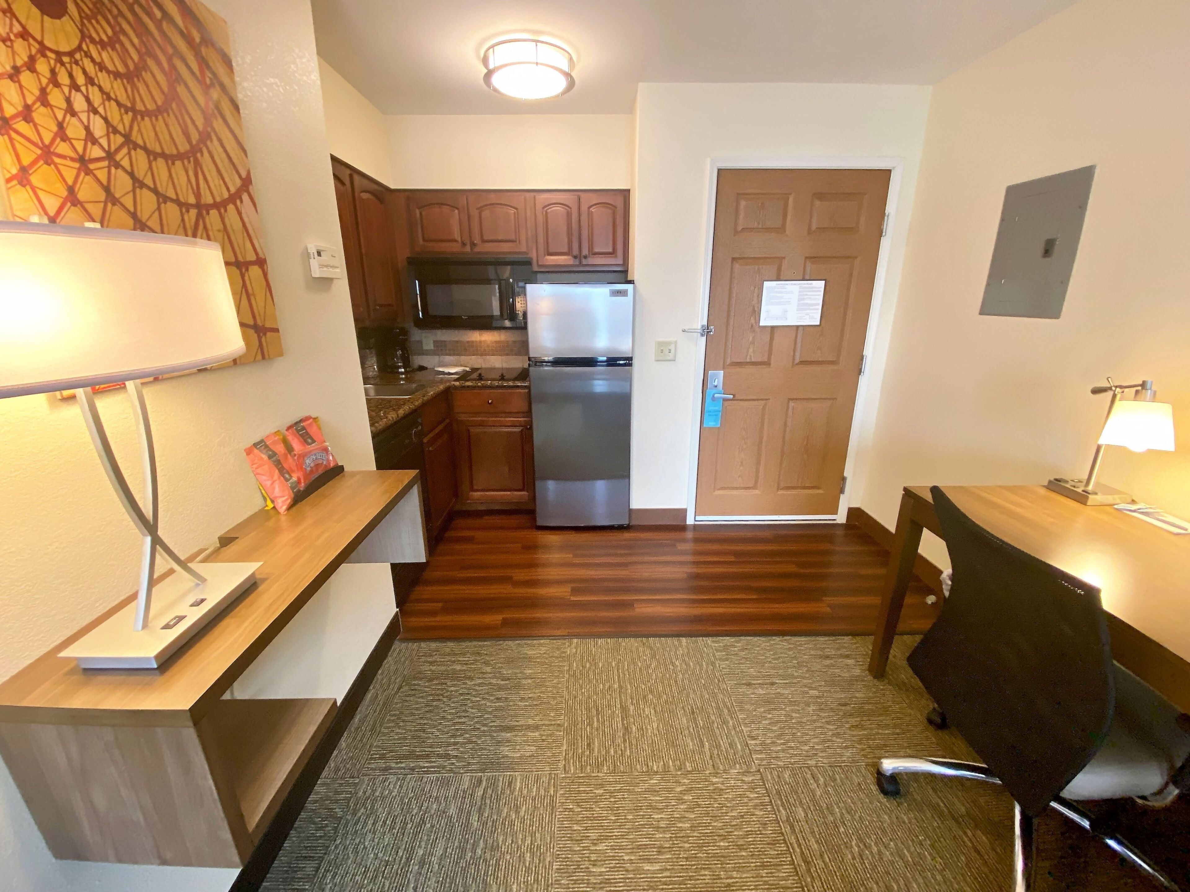 A compact kitchen with a refrigerator, microwave, and stove. There is a wooden desk with a lamp and a chair. The room has hardwood flooring and a carpeted area.