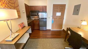 A compact kitchen with a refrigerator, microwave, and stove. There is a wooden desk with a lamp and a chair. The room has hardwood flooring and a carpeted area.
