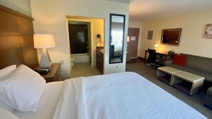 A hotel room with a bed, a desk, a chair, a sofa, a mirror, and a bathroom visible through an open door. - Great Falls Studio Suite | Free Breakfast! (Great Falls)