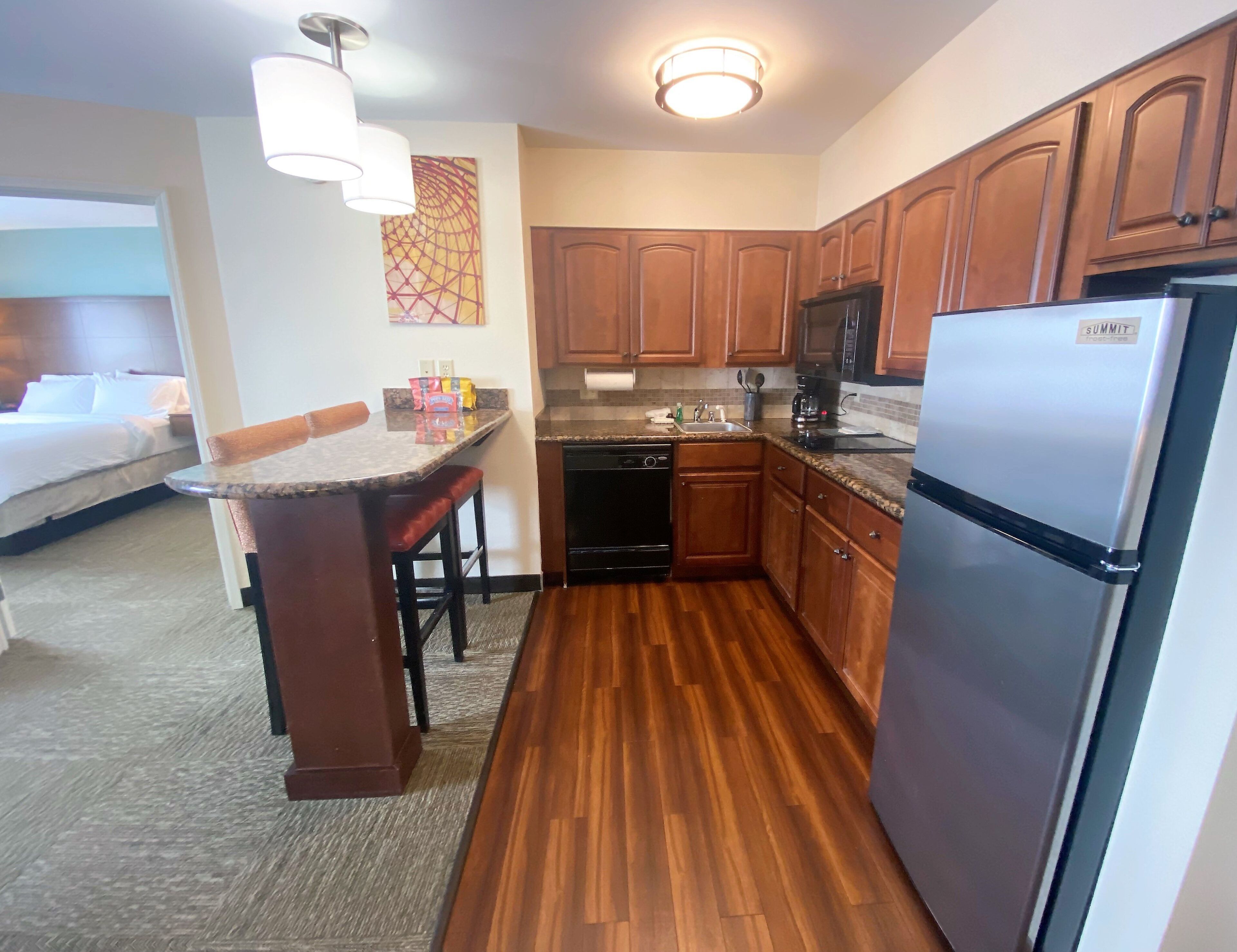 A compact hotel kitchen with wooden cabinets, a stainless steel refrigerator, a dishwasher, and a small bar area with a marble countertop.