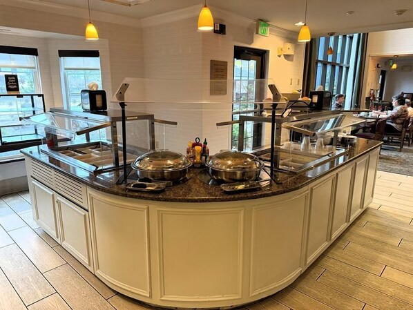 Un espace buffet doté d’un comptoir incurvé, de plats de service en acier inoxydable et d’un éclairage jaune suspendu au plafond.