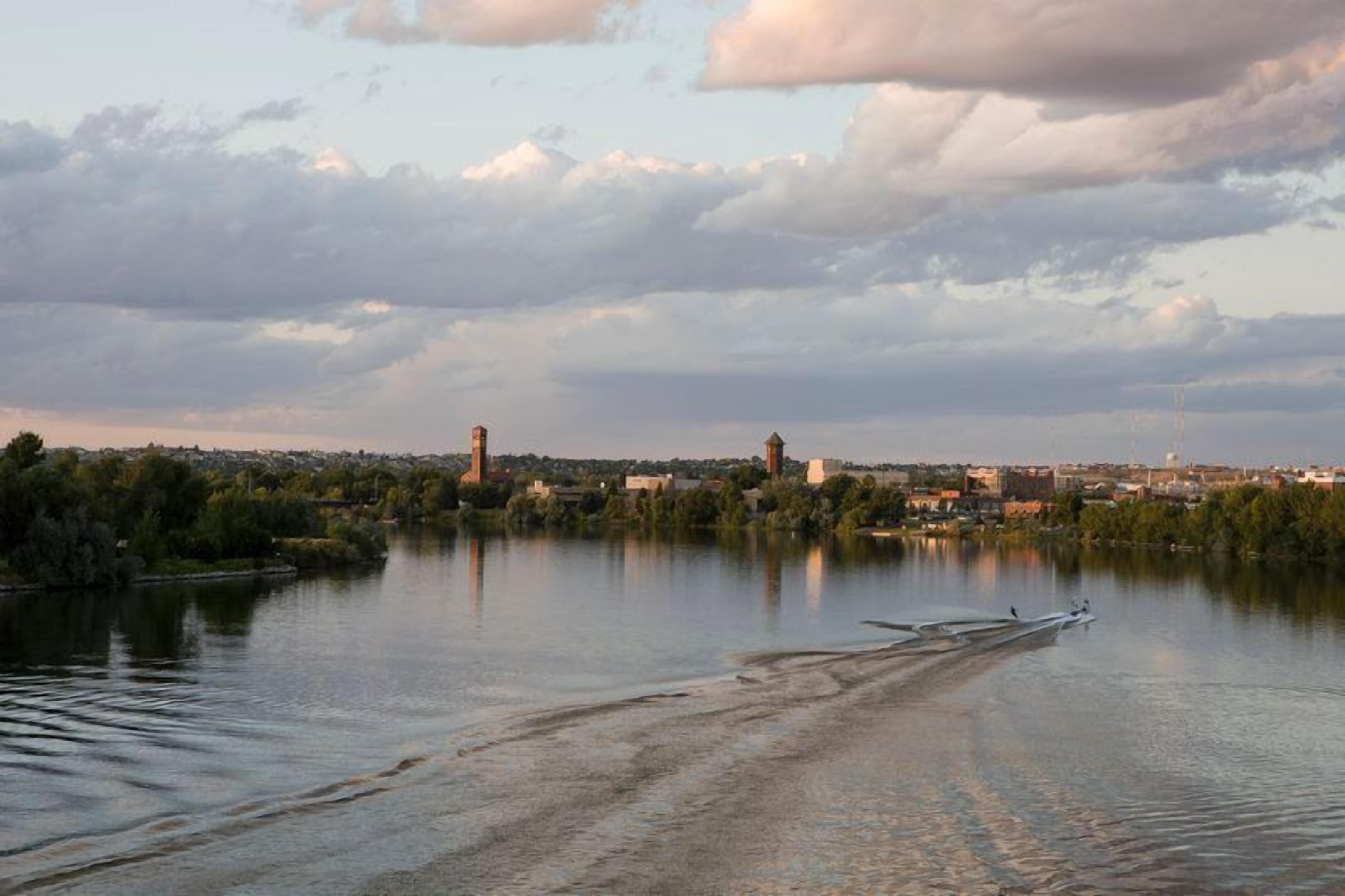 Une rivière paisible où flottent des bateaux, une vue lointaine sur la ville et un ciel nuageux.