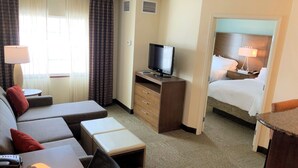 A hotel room with a sofa, a TV on a wooden stand, a bed with white linens, and a small table with a lamp. - Missouri River View Suite | Free Breakfast! (Great Falls)