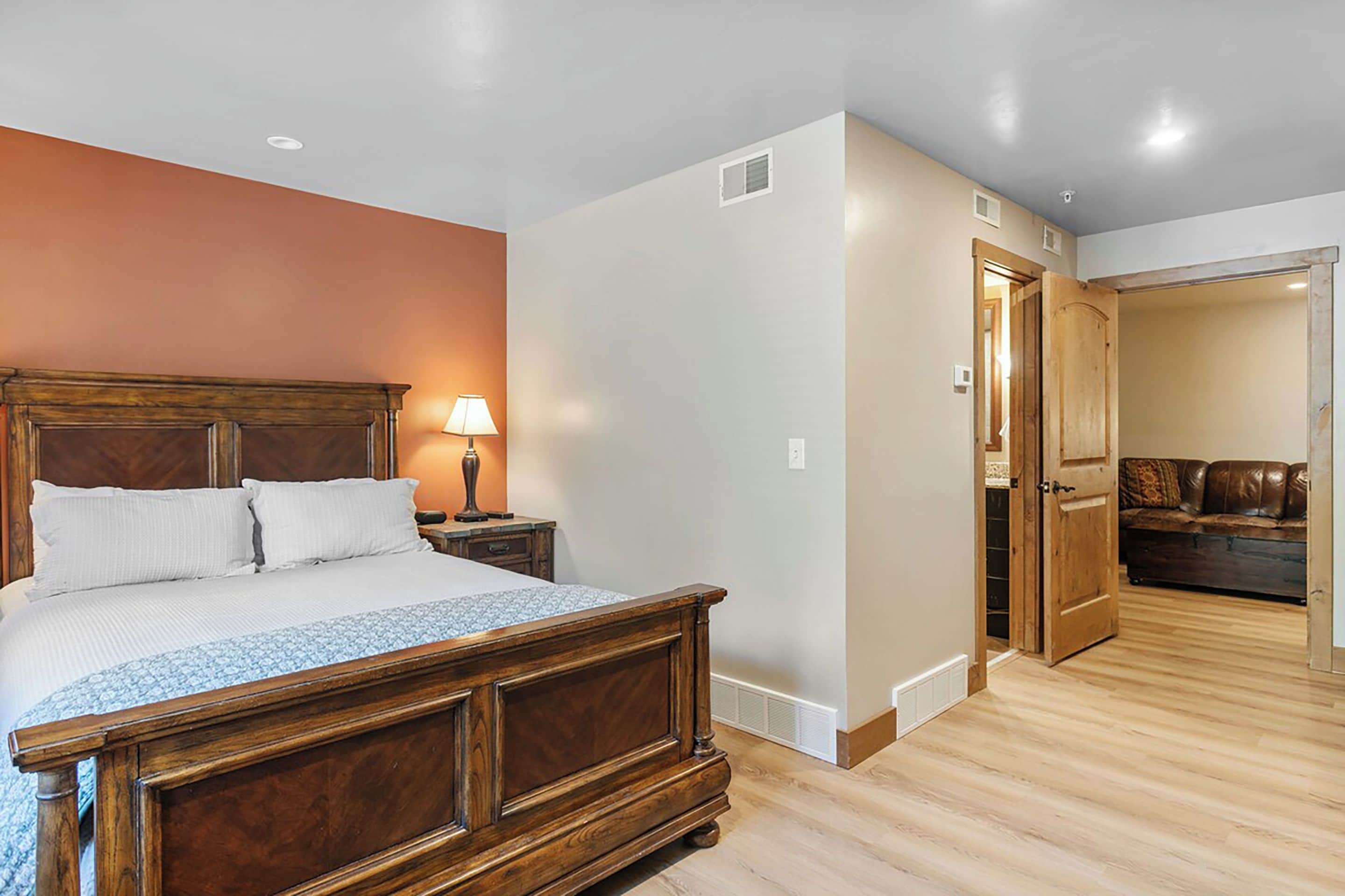 A bedroom with a wooden bed, a nightstand with a lamp, and a door leading to a living area with a couch.