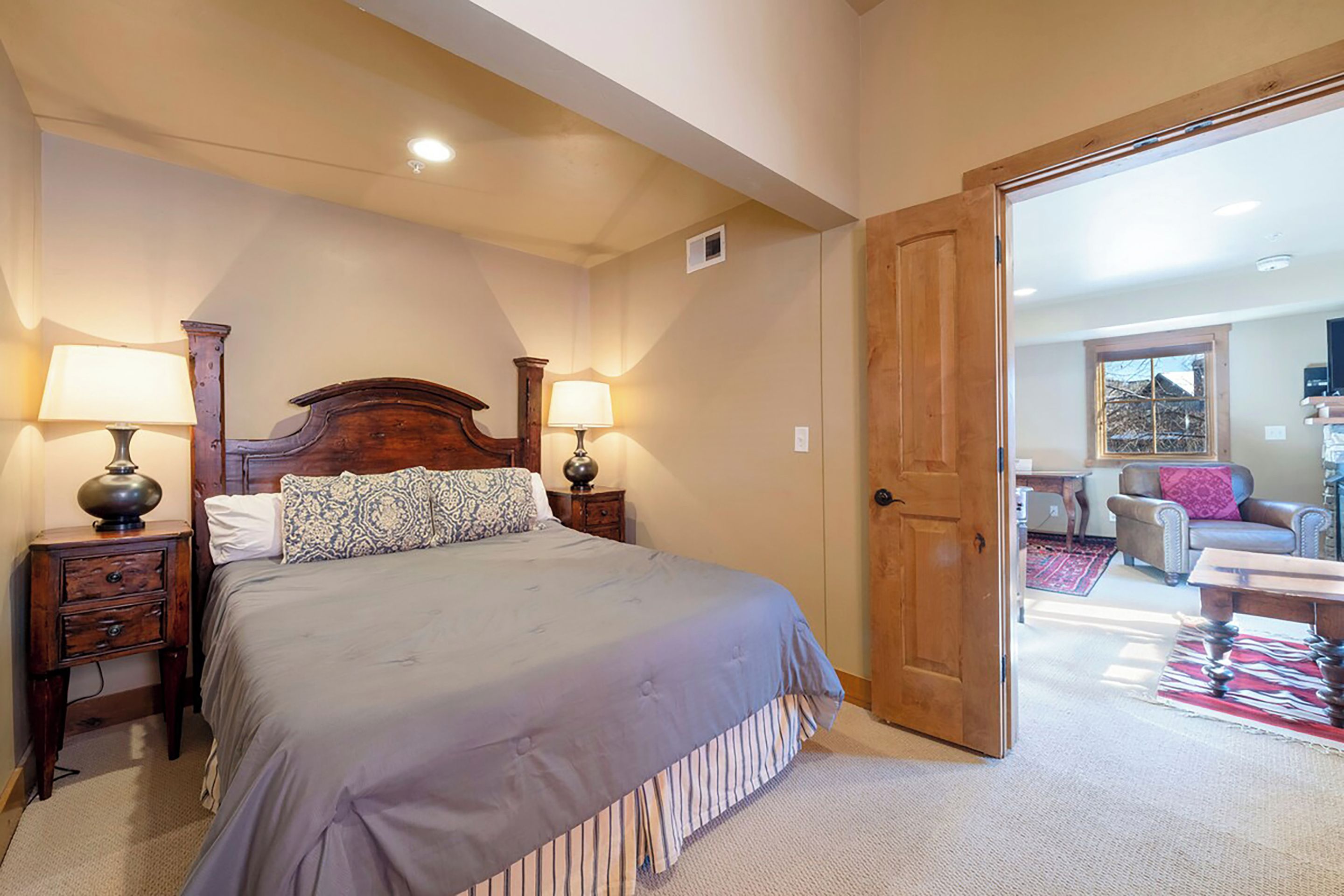 Quarto com uma cama grande, duas mesas de cabeceira com luminárias, uma porta de madeira e vista para uma sala de estar com sofá e mesa.