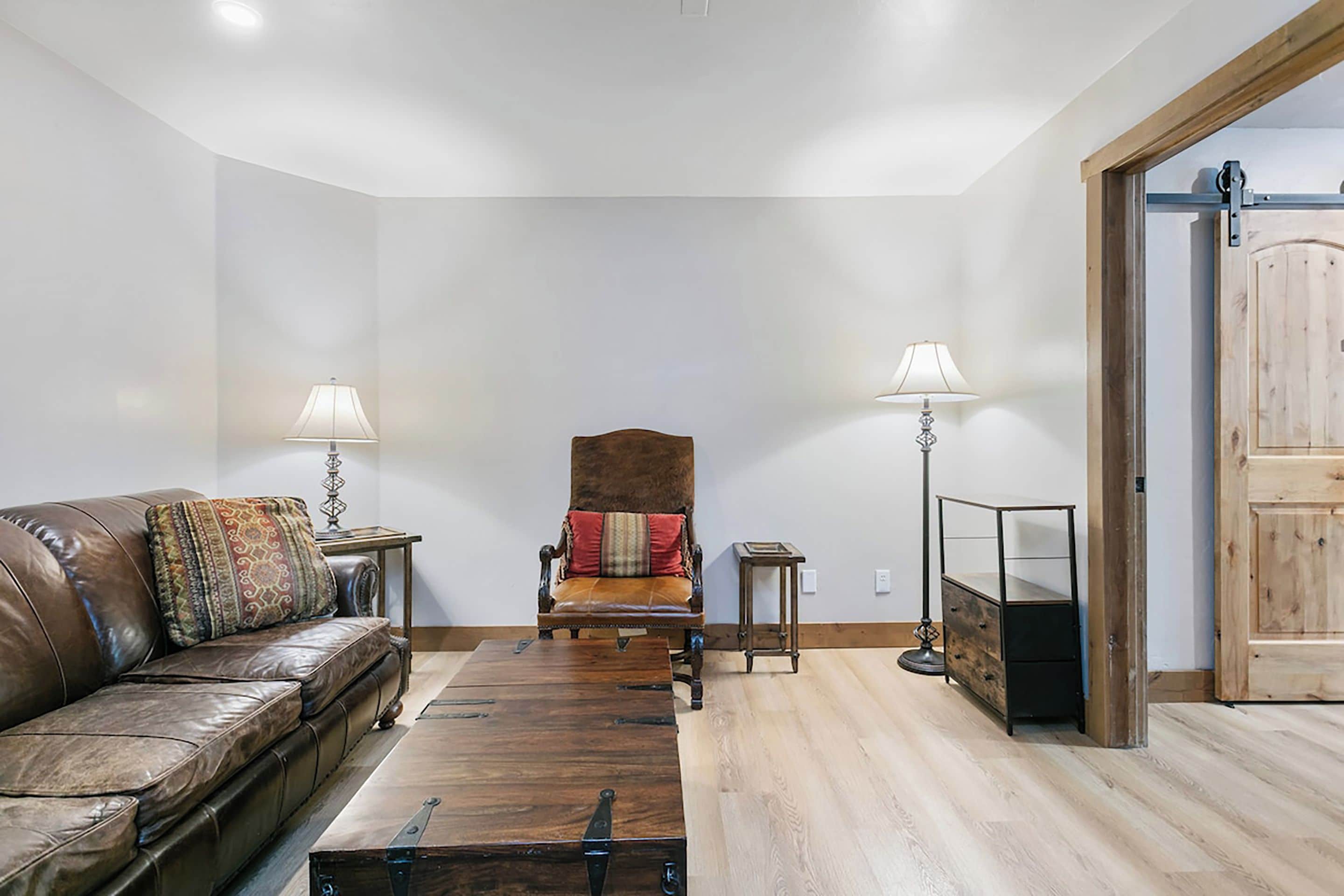 A living room with a leather sofa, a wooden coffee table, a brown armchair with a red cushion, two floor lamps, and a wooden cabinet.