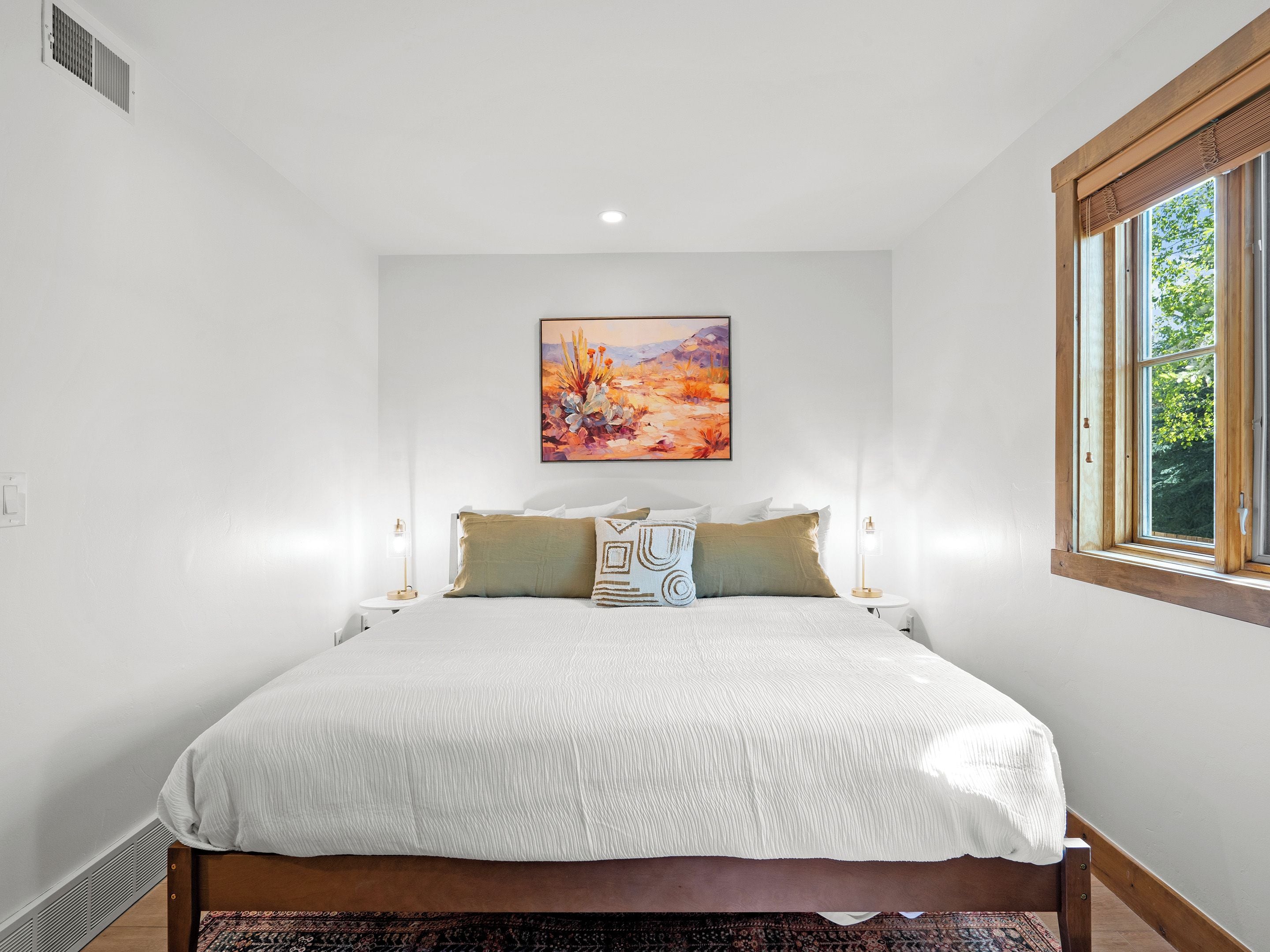 A bedroom with a large bed, a painting on the wall, and a window with a view of greenery.