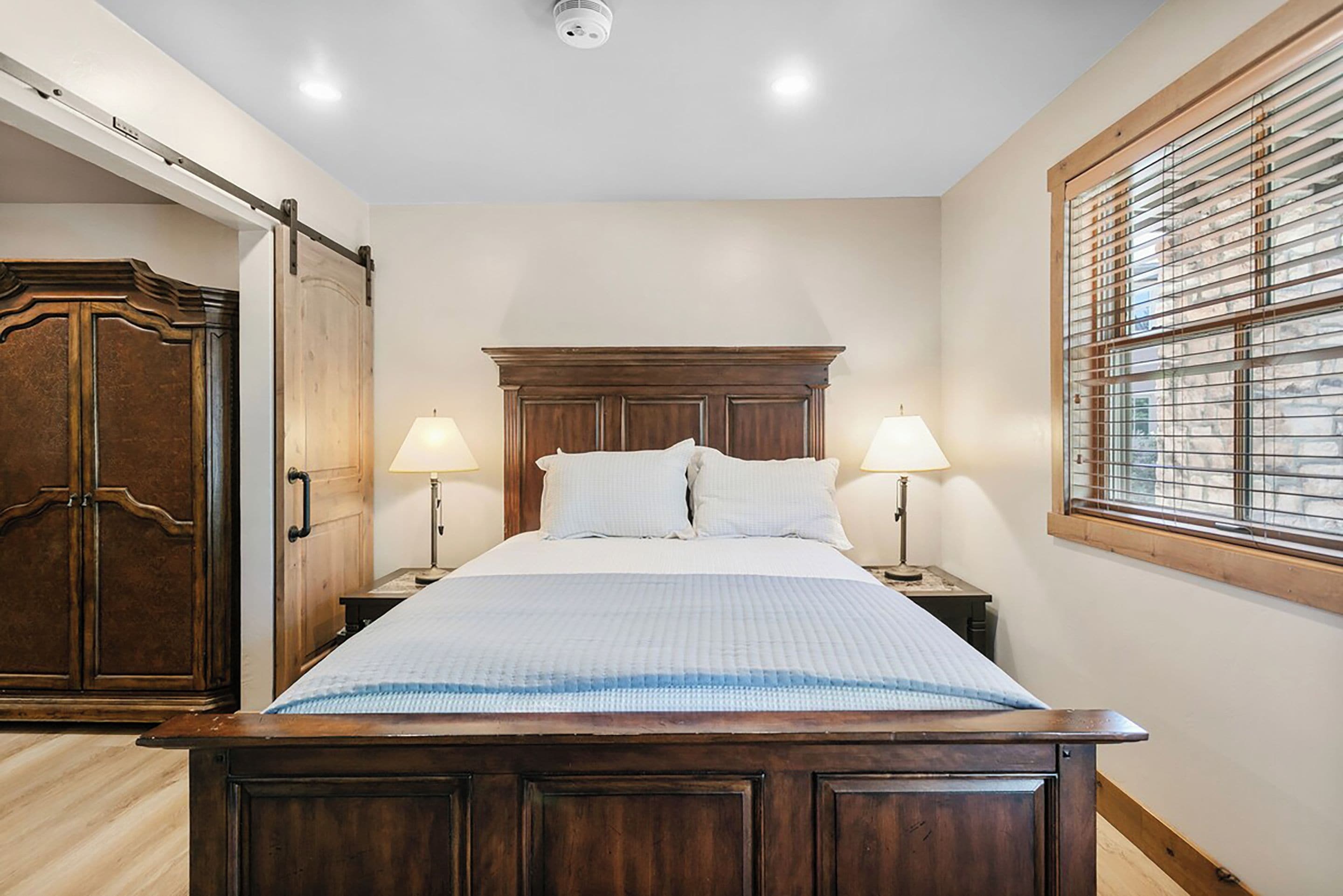 A bedroom with a wooden bed, two bedside lamps, a window with blinds, and a ceiling light.
