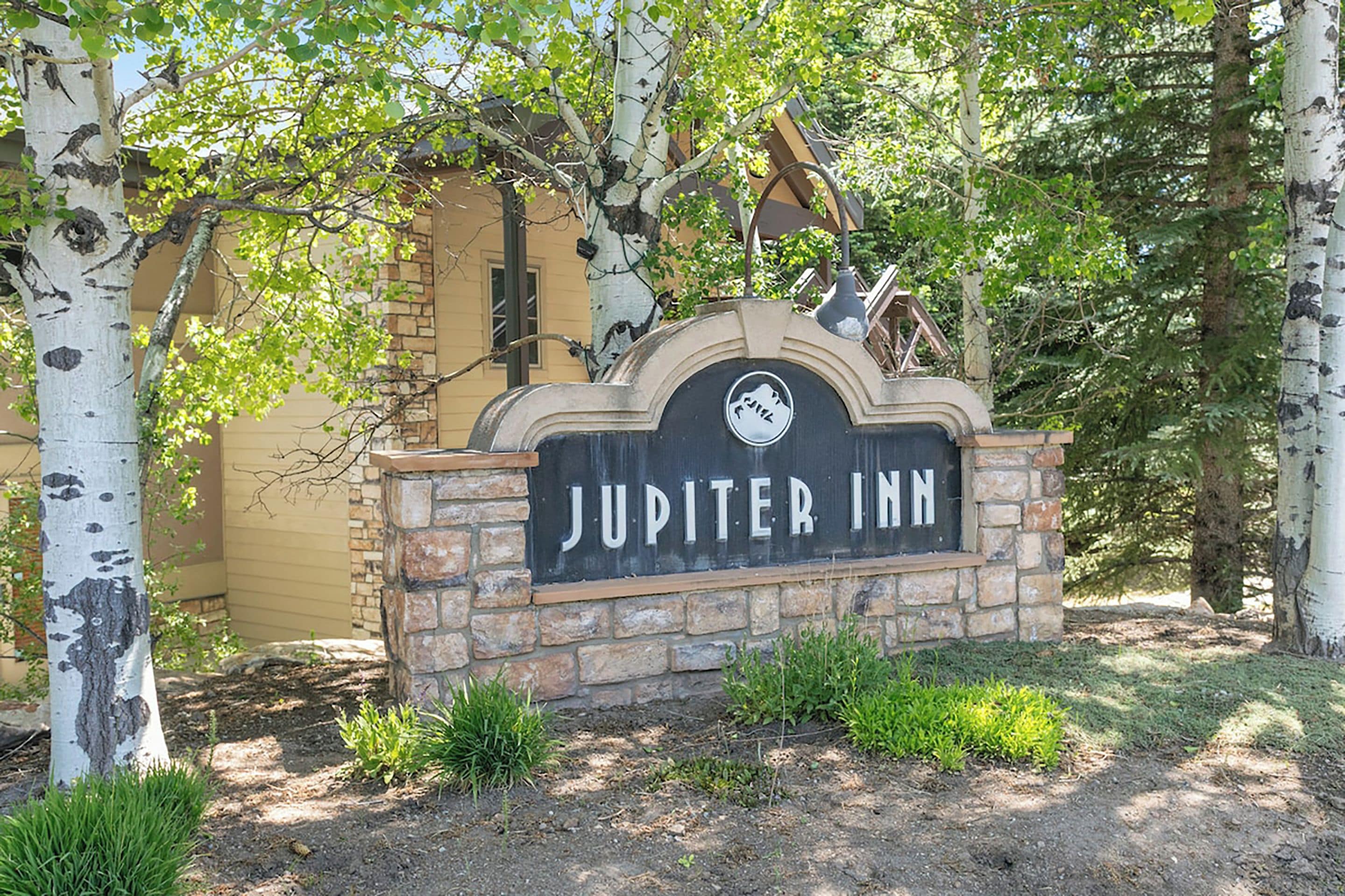 Placa do Jupiter Inn, com base de pedra e topo metálico, cercada por árvores e vegetação.
