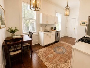 A kitchen with white cabinets, a wooden dining table, a rug, and pendant lighting. - The Gilded Magnolia | Historic Savannah Stay (Savannah)