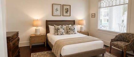 Un dormitorio con una cama grande, dos lámparas de noche, una mesita de noche de madera, una silla, una ventana con persianas y dos cuadros en la pared.