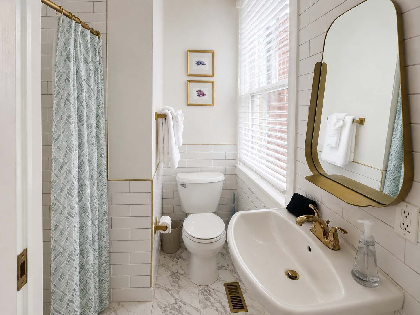 A bathroom with a toilet, sink, and shower curtain. There are framed pictures on the wall and a window with blinds.