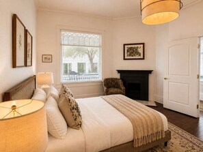 A bedroom with a bed, a chair, a fireplace, and a window with blinds. - The Gilded Magnolia | Historic Savannah Stay (Savannah)