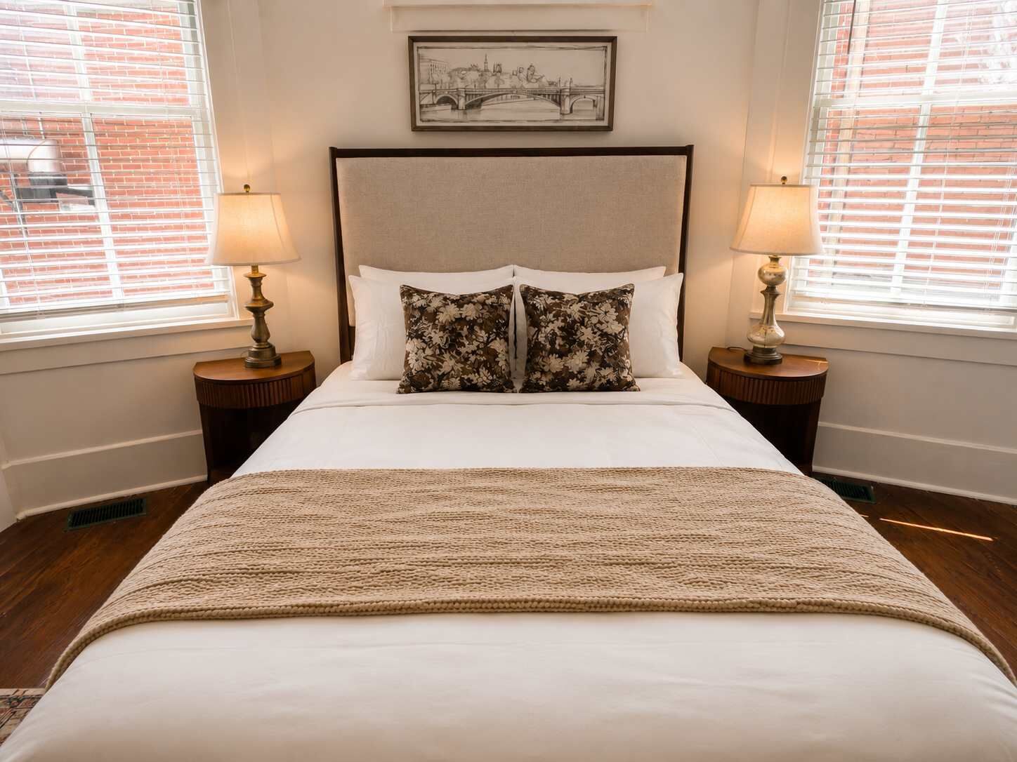 Quarto de hotel com uma cama grande, duas mesinhas de cabeceira, duas luminárias, um quadro na parede e uma janela com persianas.