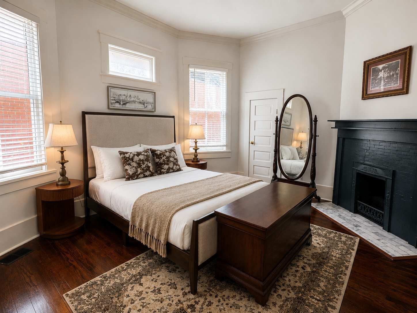 A bedroom with a bed, bedside tables, a dresser, a mirror, a fireplace, and two windows with blinds.