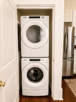 A compact laundry room with a top-loading washer and a front-loading dryer. - The Gilded Magnolia | Historic Savannah Stay (Savannah)