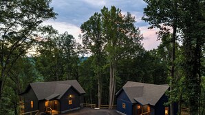 A cabin with a wooden deck and a paved driveway, surrounded by dense forest. - NEW CABIN: Sauna | Hot Tub | Fire Pit | EV (Gatlinburg)