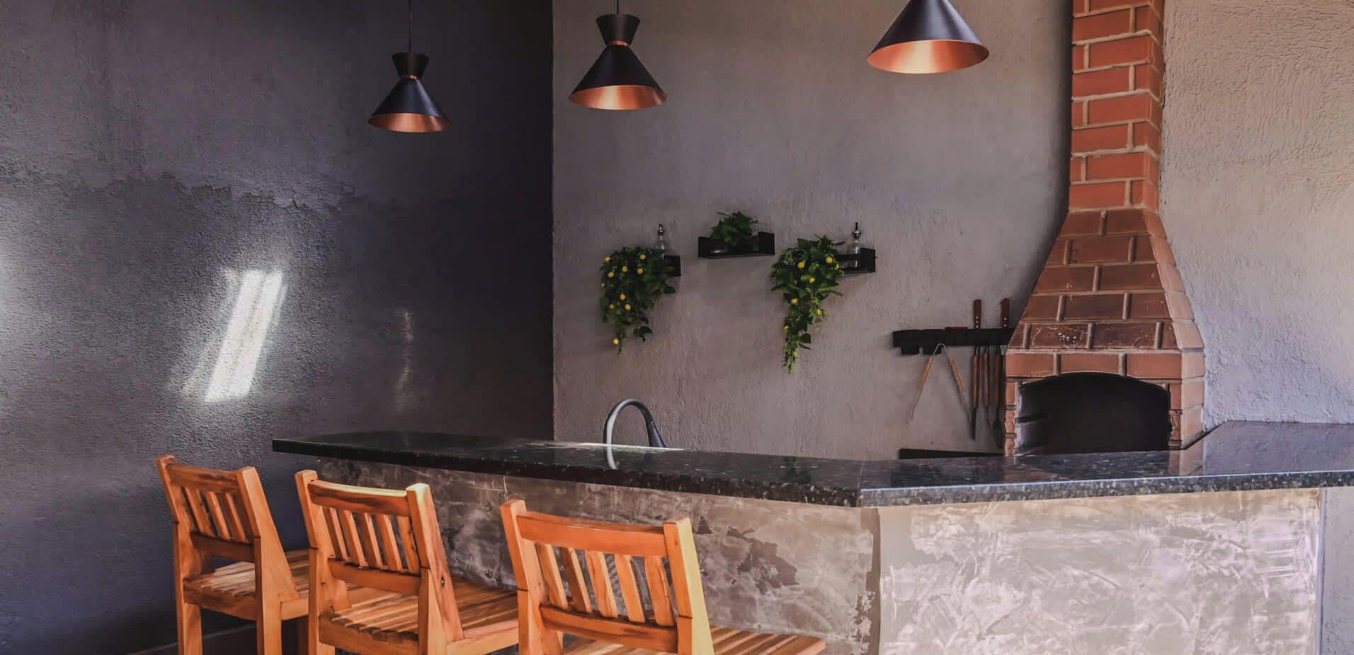 A kitchen with a dark wall, a brick oven, wooden chairs, and pendant lights.