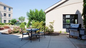A patio with a table and chairs, a grill, and a building in the background. - Accessible 1BR Suite | Free Daily Breakfast & Pool (Madison)