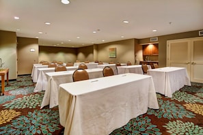 A conference room with multiple tables and chairs, carpeted floor, and a kitchenette area.