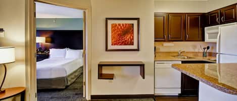 A hotel room with a bed, a desk, a lamp, a framed picture, a kitchen area with cabinets and a microwave, and a carpeted floor.