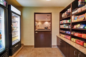 A hotel pantry with a refrigerator stocked with beverages, a microwave, and shelves with snacks and condiments.