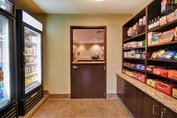 A hotel pantry with a refrigerator stocked with beverages, a microwave, and shelves with snacks and condiments.