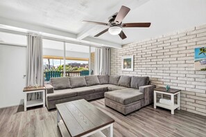 A modern living room with a sectional sofa, a wooden coffee table, a ceiling fan, and a brick wall. - Jade’s Getaway | 2BR Condo Near the Beach with Pool & Private Boardwalk (Port Aransas)