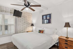 Ein Schlafzimmer mit einem Bett, einem Deckenventilator, einem Fenster mit Jalousien, einer Ziegelwand und einem gerahmten Bild an der Wand.