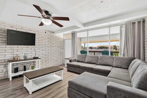 A modern living room with a flat-screen TV, a ceiling fan, a sofa, and a large window with curtains. - Jade’s Getaway | 2BR Condo Near the Beach with Pool & Private Boardwalk (Port Aransas)