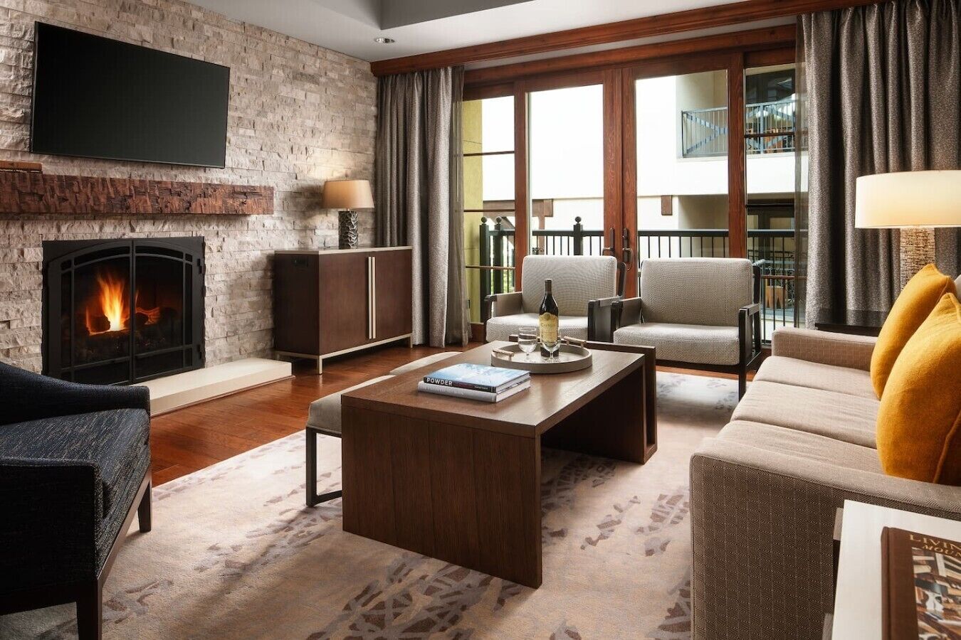 A living room with a fireplace, a flat-screen TV mounted on a brick wall, a sofa, a coffee table with a book and a bottle, and a view of the outdoors through large windows.