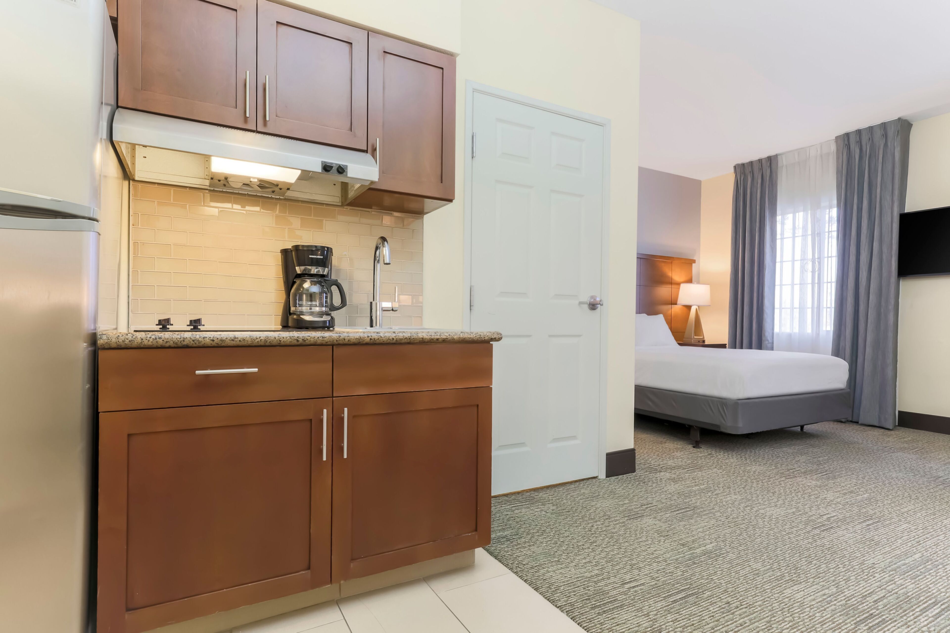 Habitación de hotel con kitchenette, una cama, una mesita de noche con lámpara y una ventana con cortinas.