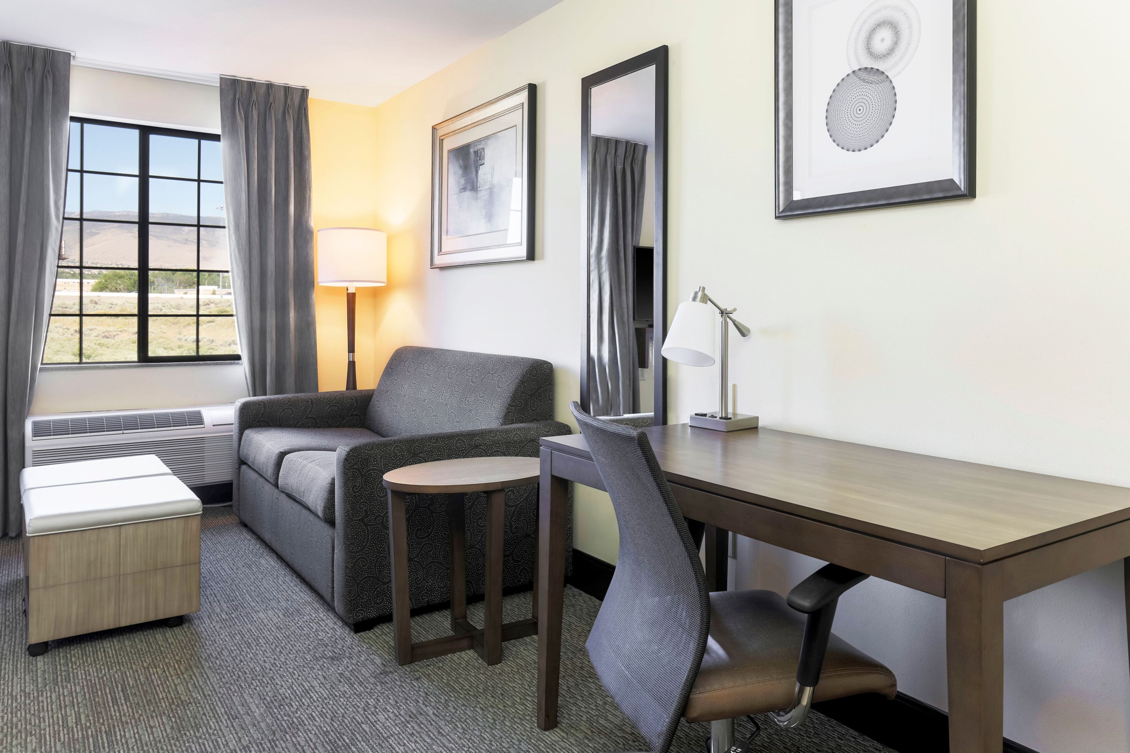Ein Hotelzimmer mit einer Couch, einem kleinen Tisch, einem Schreibtisch, einem Stuhl, einer Lampe, einem Bett und einem Fenster mit Vorhängen.