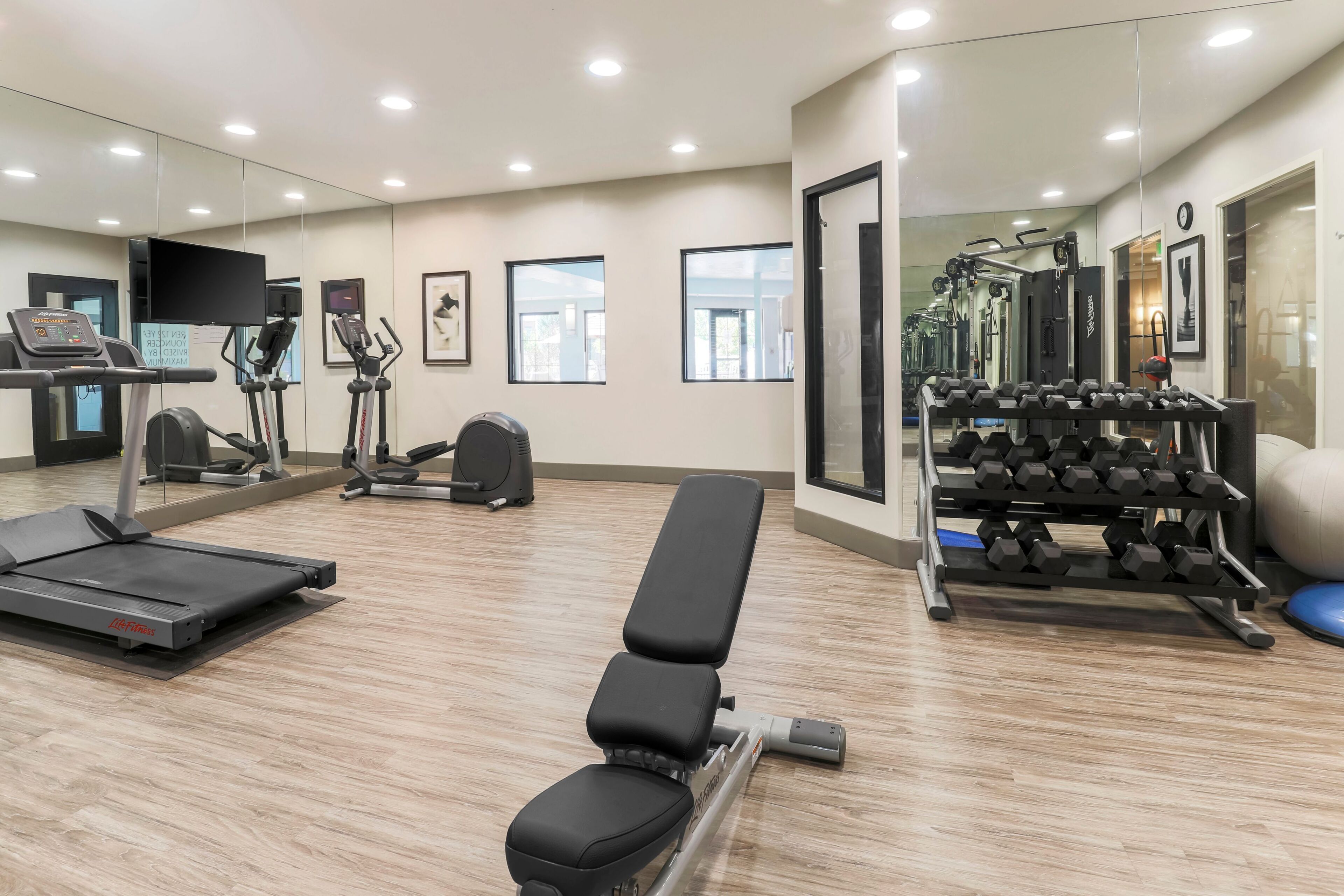 Uma academia com equipamentos de exercícios, esteira, banco e parede com espelhos.