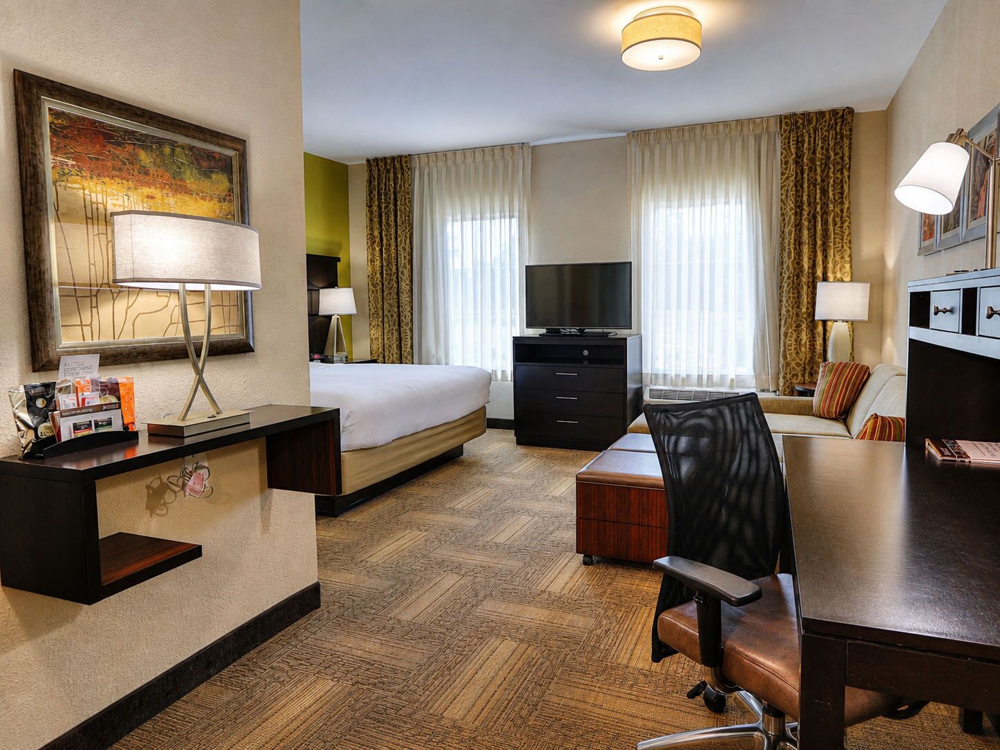 Quarto de hotel com cama, escrivaninha, cadeira, televisão e um quadro na parede.