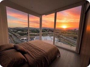 Quarto com uma cama grande e vista para um pôr do sol sobre a paisagem urbana.