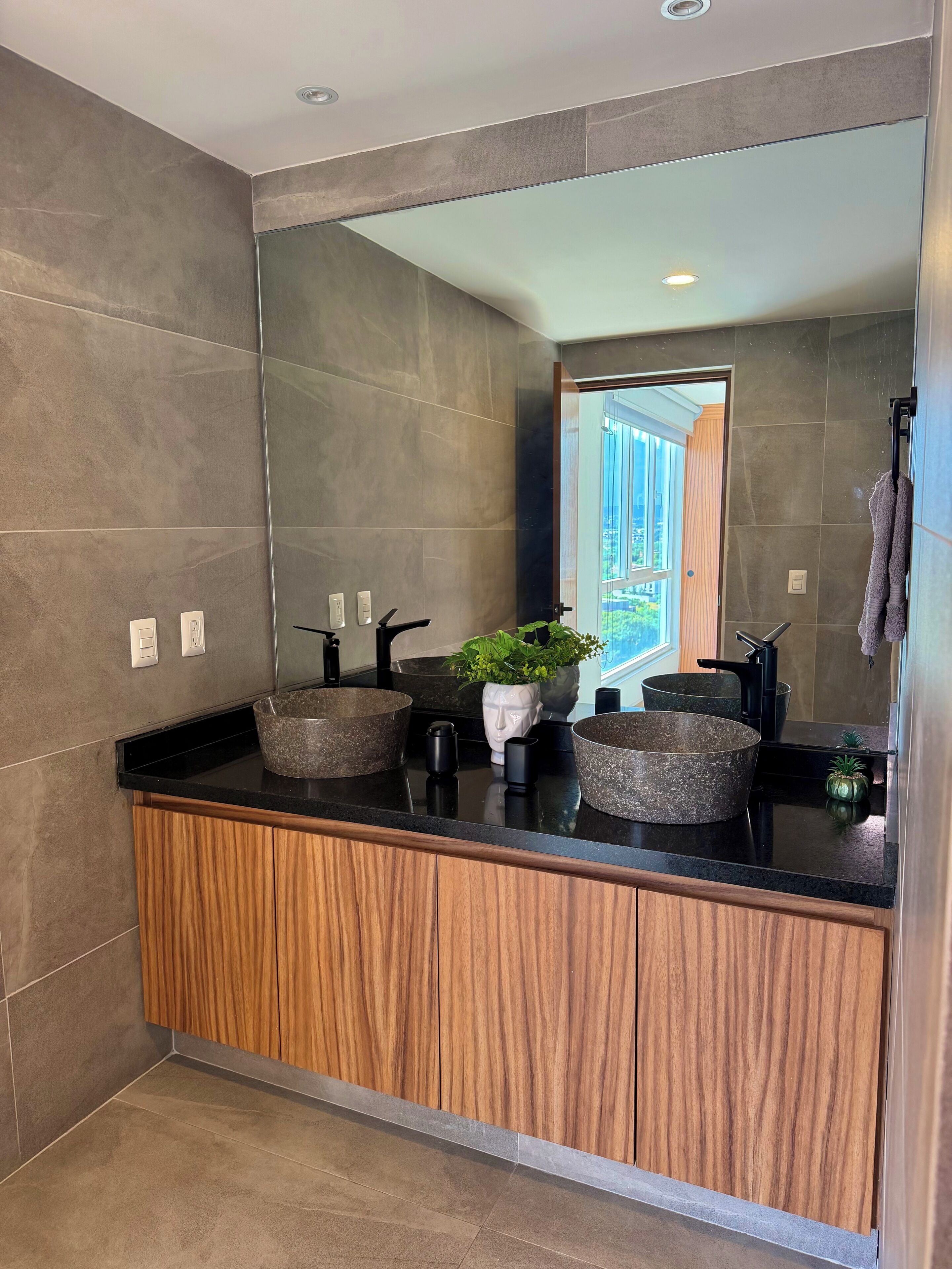 A modern bathroom with a large mirror, two sinks with stone basins, wooden cabinetry, and a potted plant.