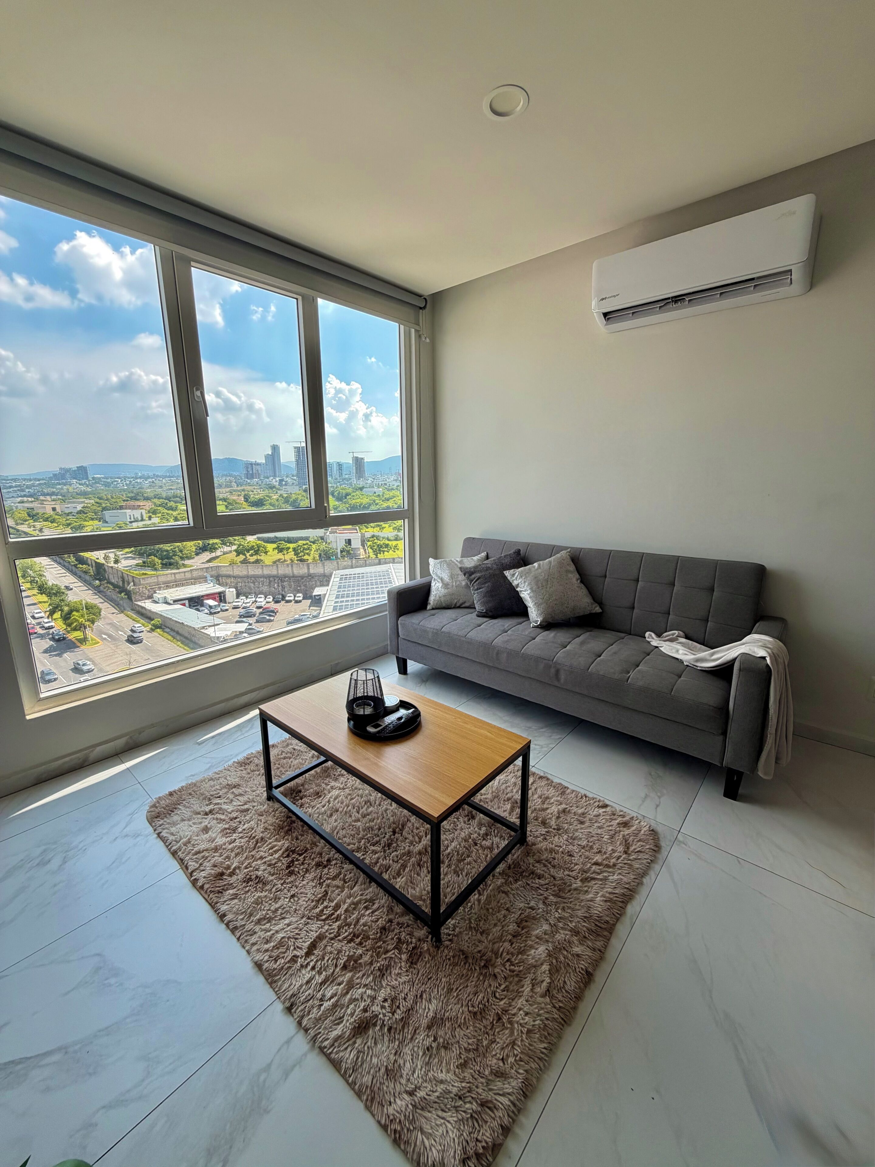 Una sala de estar moderna con un sofá gris, una mesa de centro de madera y un ventanal con vistas a la ciudad.