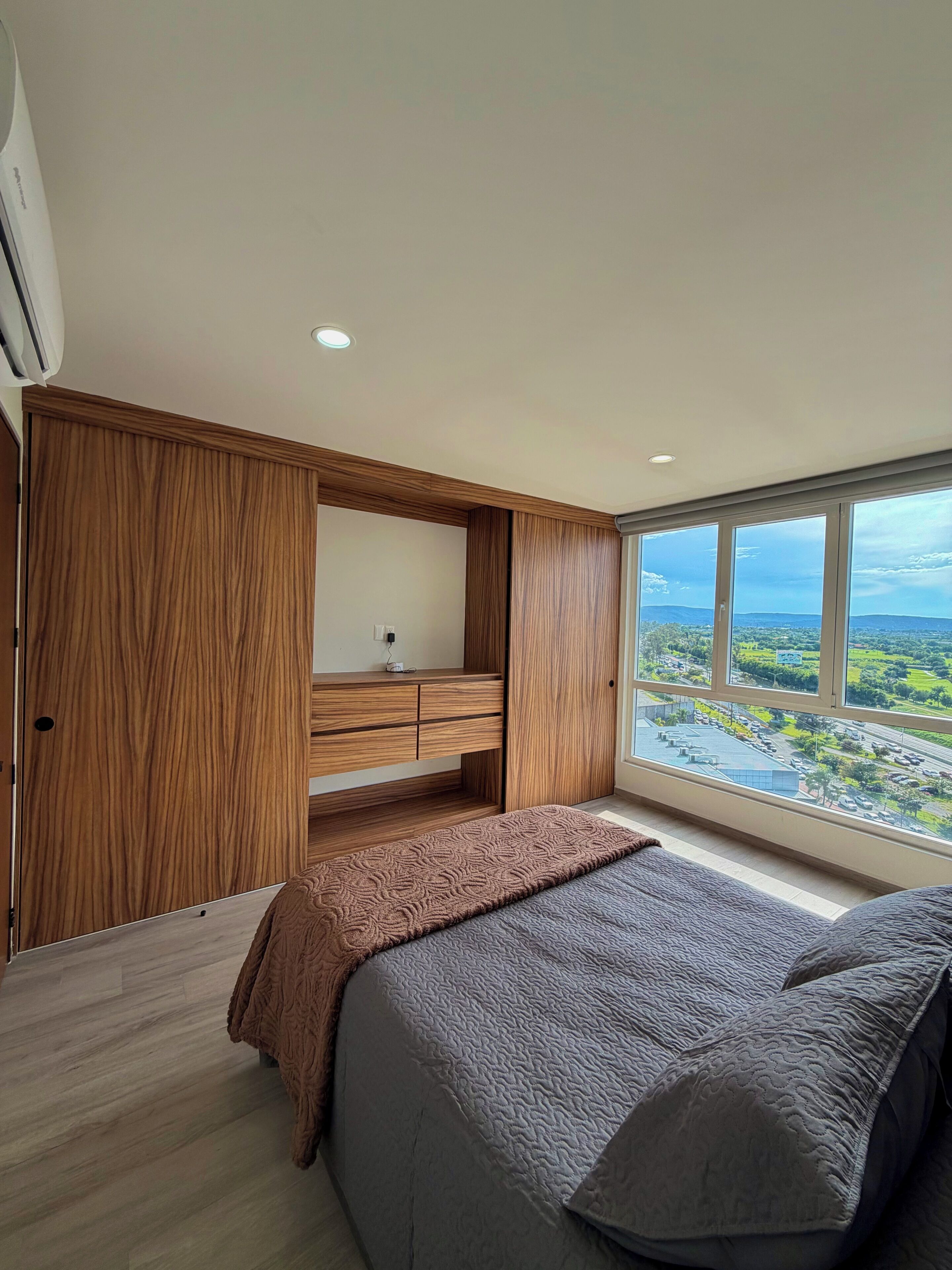 Ein Schlafzimmer mit großem Fenster, eingebautem Kleiderschrank und einem Bett mit grauer Tagesdecke.