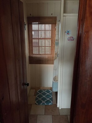 Ein schmaler Flur mit einer Holztür links, einem Fenster mit Jalousien, einem kleinen Teppich und einem Kühlschrank mit Aufkleber rechts.
