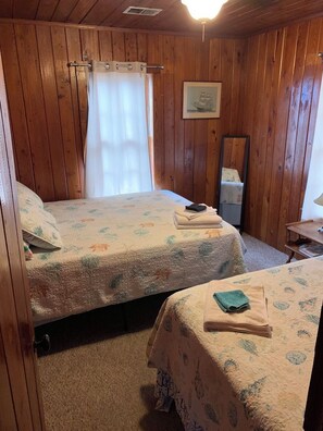 A room with wooden paneling, two beds with floral bedding, a mirror, and a window with a curtain.