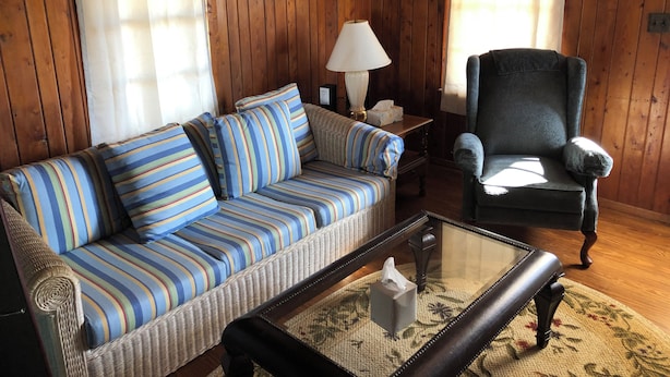 A wooden-paneled room with a striped sofa, a dark armchair, a glass coffee table, and a lamp. - 2-bedroom cottage with AC in welcoming Kitty Hawk (Kitty Hawk)