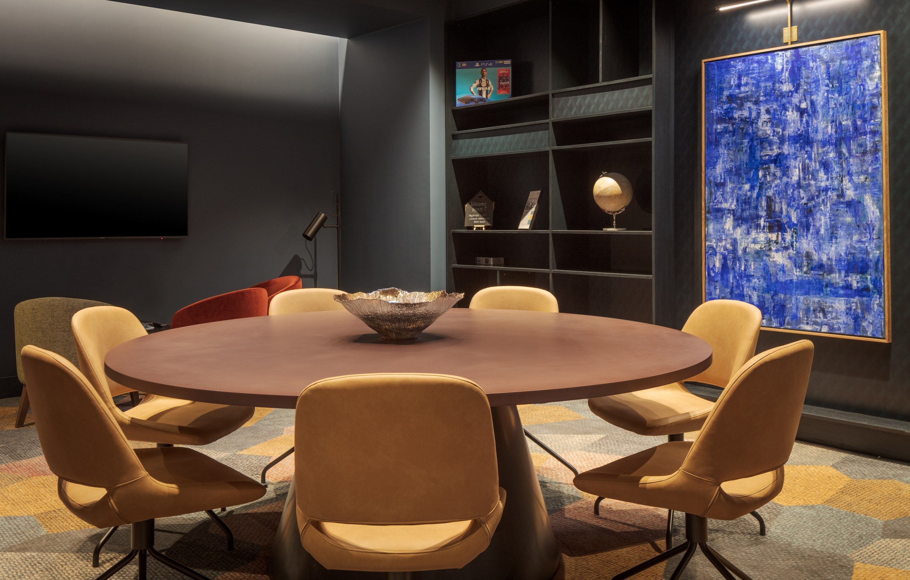A modern conference room with a round table, six chairs, a large abstract painting, and a bookshelf with awards.