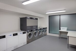 A laundry room with multiple washing machines, a dryer, a small table with a basket, and a large window with frosted glass.