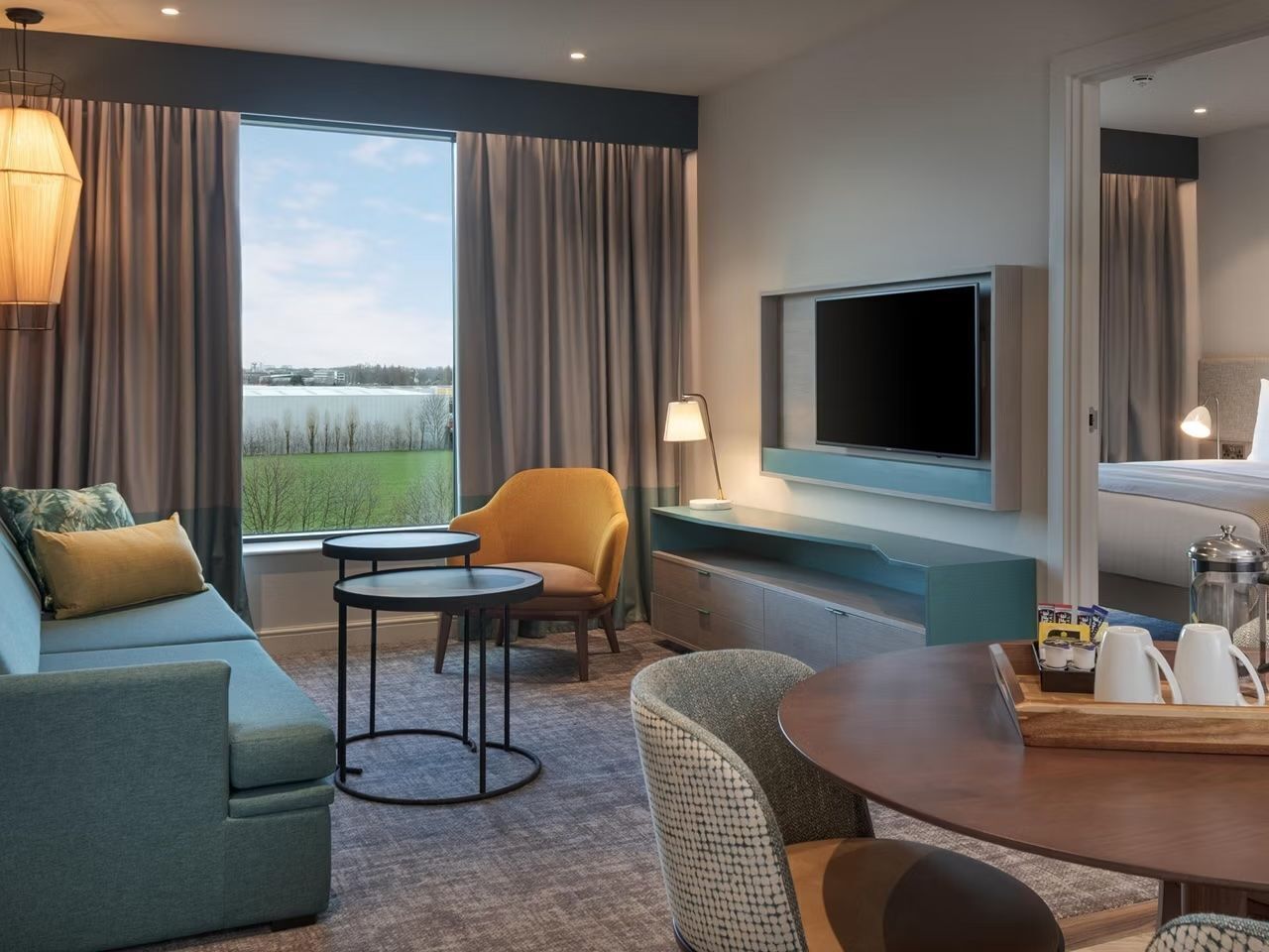 A hotel room with a sofa, armchair, TV, and a dining table with a tray of cups.